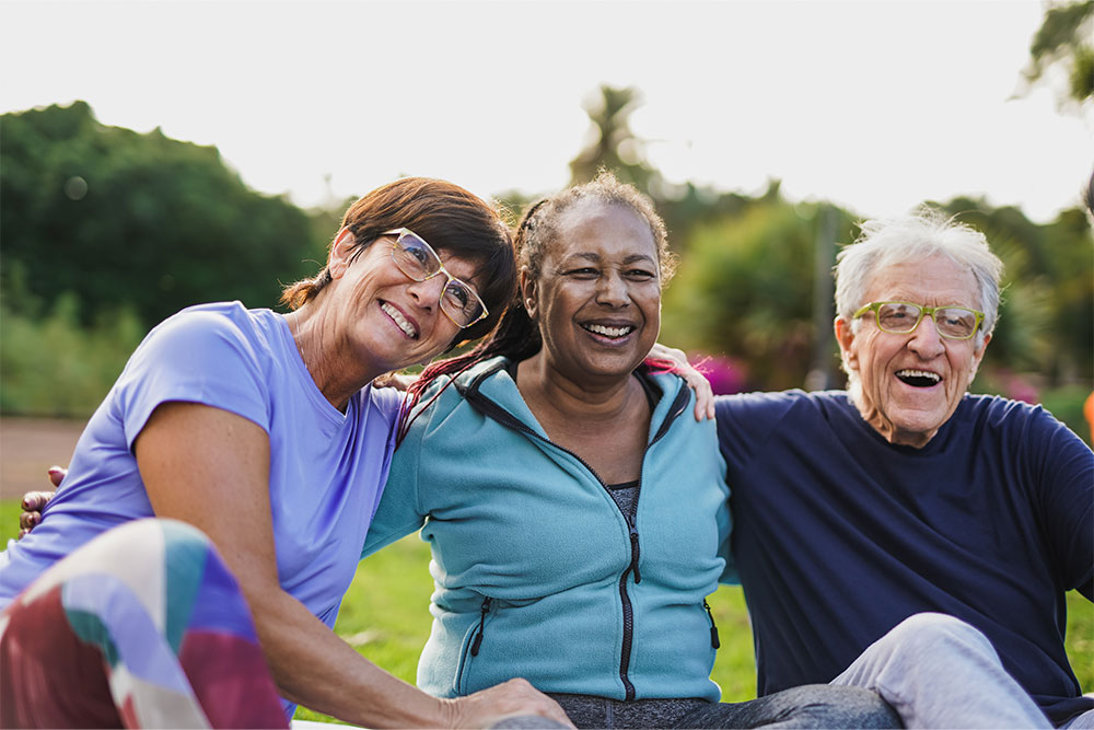 Three elderly adults sit in a park, grab each others shoulders and smile.