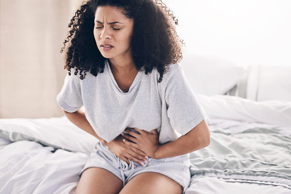 A woman sits on a bed and holds her stomach in pain.